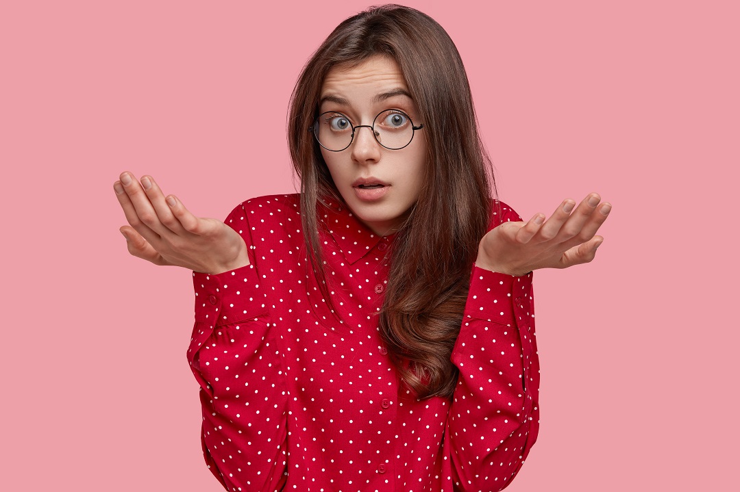No idea. Unaware careless young woman spreads hands in clueless gesture, looks doubtfully at camera, makes decision, dressed in polka dot blouse, isolated over pink studio wall, has raised arms